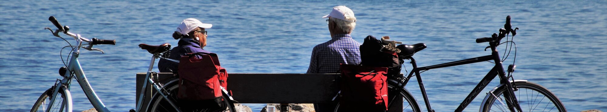 Zwei ältere Menschen haben ihre Fahrräder an einer Bank abgestellt und schauen auf den See